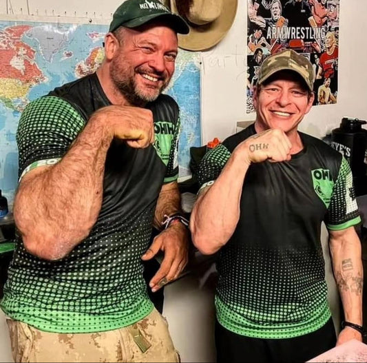Two men in matching green and black shirts flexing their muscles in front of a map and arm wrestling poster.