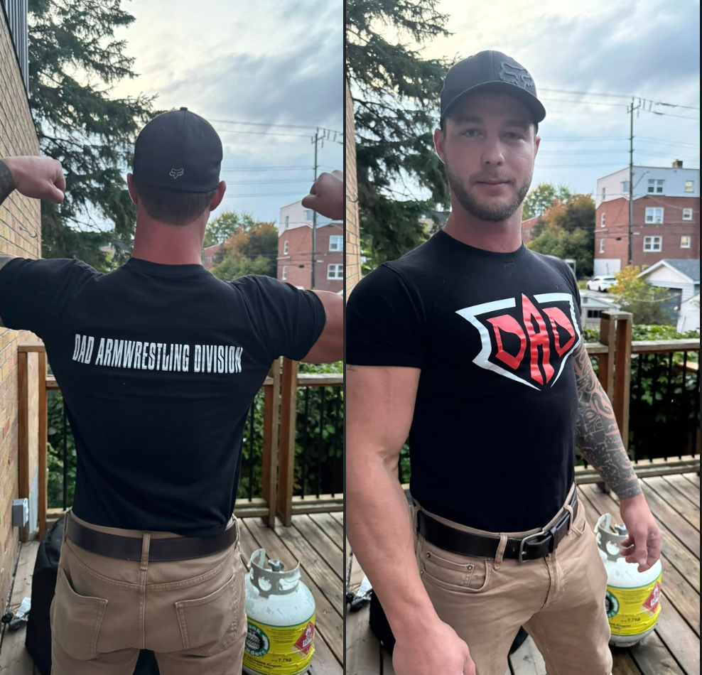 Man wearing a black t-shirt with 'Dad Armwrestling Division' on the front and back, standing on a deck.